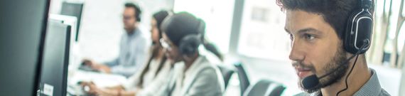 four people sit in front of computer screens with headphone and microphone headsets on